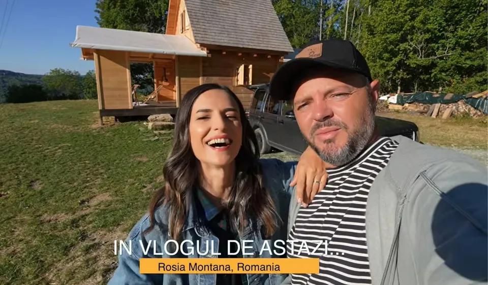 Un bărbat cu șapcă și o femeie zâmbesc în prim-plan, având o casă din lemn și un autoturism în fundal pe un teren verde.