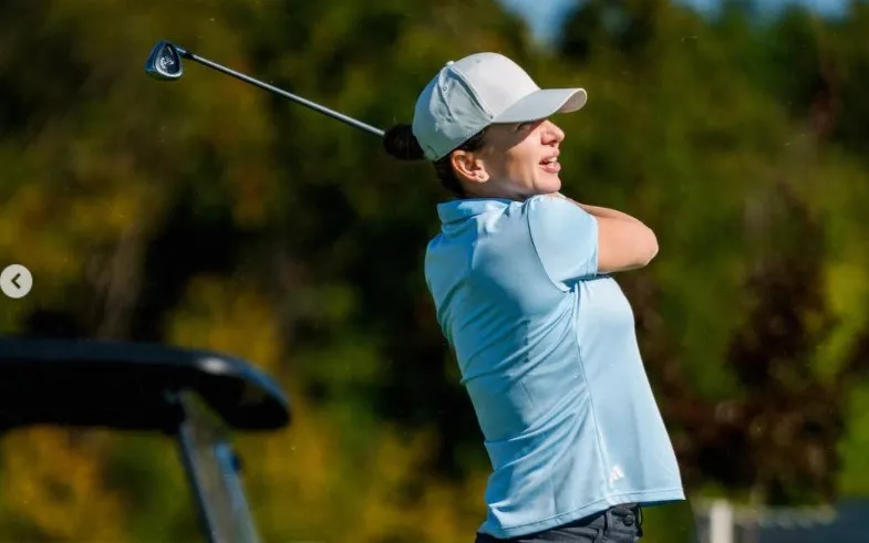 O femeie cu șapcă albă și tricou bleu ține un stick de golf în poziție de final de lovitură pe un teren verde.