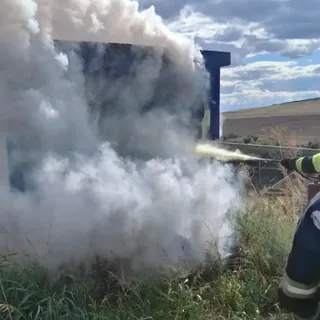 Un jet de apă este direcționat spre un obiect mare acoperit de fum alb dens într-un câmp cu iarbă înaltă.