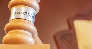 Un ciocan de lemn cu un inel metalic pe care scrie textul parțial vizibil CONSTITUTION se află pe o suprafață lemnoasă.