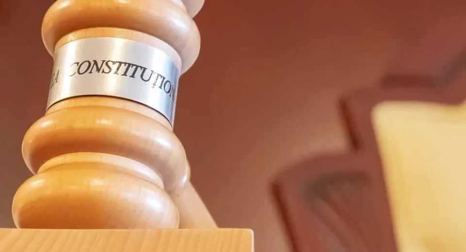 Un ciocan de lemn cu un inel metalic pe care scrie textul parțial vizibil CONSTITUTION se află pe o suprafață lemnoasă.