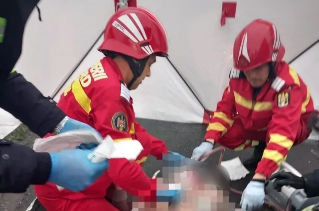 Două persoane în uniforme roșii cu benzi galbene și căști de protecție roșii efectuează manevre medicale asupra unei persoane întinse pe sol.