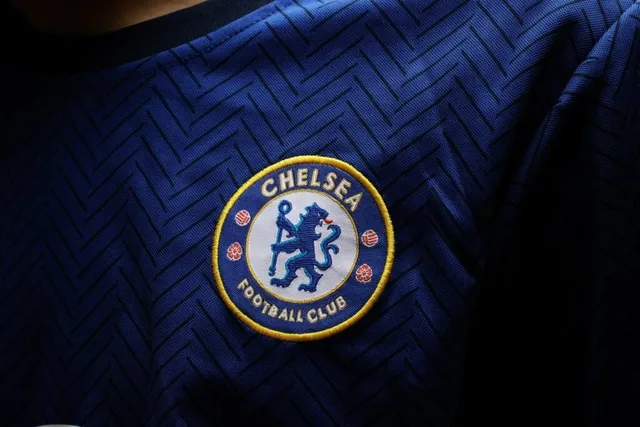 Un logo circular albastru și galben cu un leu și textul Chelsea Football Club este cusut pe un tricou albastru cu model în zig-zag.