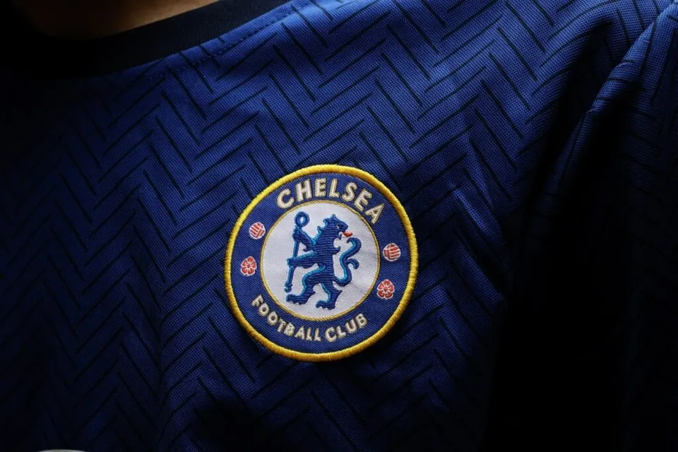 Un logo circular albastru și galben cu un leu și textul Chelsea Football Club este cusut pe un tricou albastru cu model în zig-zag.