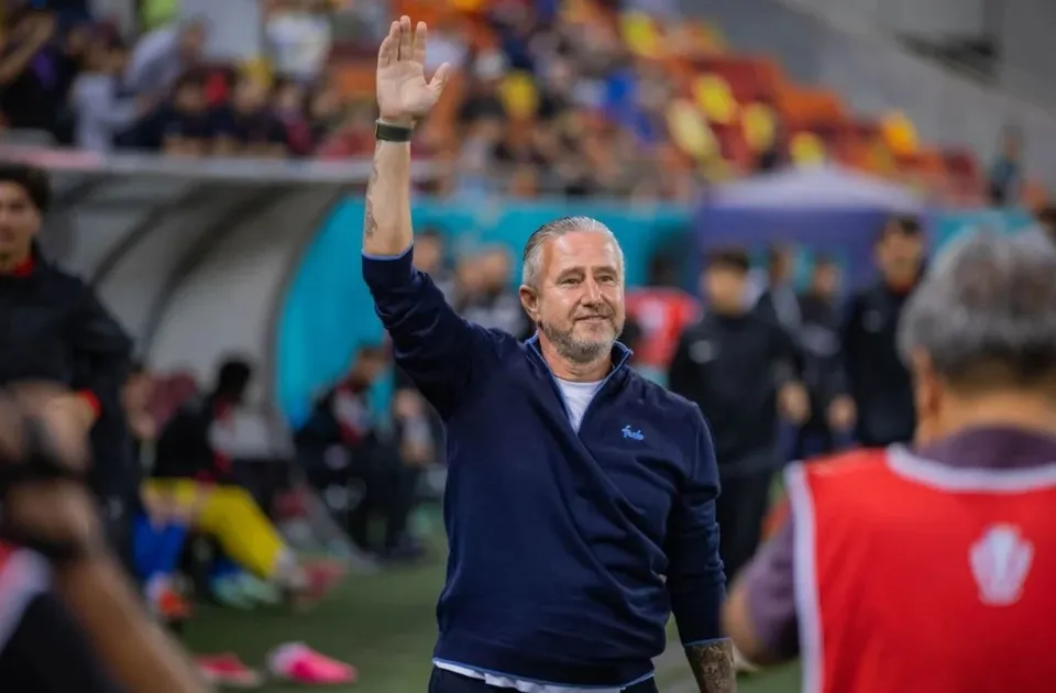 Un bărbat cu părul cărunt și bluza albastră ridică mâna dreaptă în semn de salut pe un teren de fotbal.