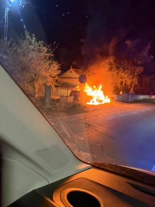 Flăcări mari ard printre copaci pe marginea unui drum asfaltat, fiind vizualizate de pe interiorul unui autoturism noaptea.