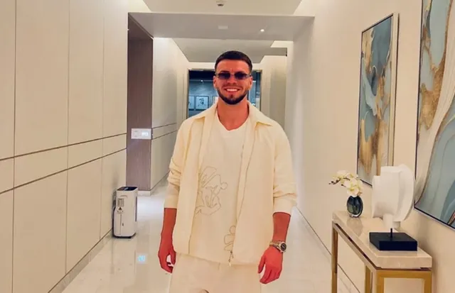 Un bărbat cu ochelari de soare și cămașă deschisă de culoare deschisă stă în picioare într-un hol cu tablouri pe perete.