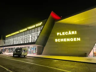 O clădire de aeroport iluminată noaptea prezintă inscripțiiile "PLECĂRI SCHENGEN" și un vehicul mic alb parcat în fața terminalului.