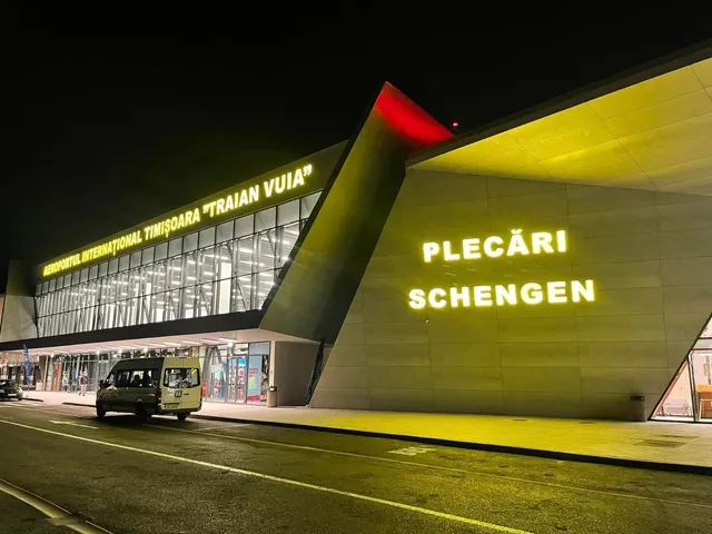 O clădire de aeroport iluminată noaptea prezintă inscripțiiile "PLECĂRI SCHENGEN" și un vehicul mic alb parcat în fața terminalului.