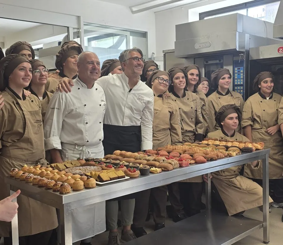 Un grup de persoane îmbrăcate în uniforme de bucătar stau în spatele unei mese metalice pe care sunt așezate diverse produse de patiserie.