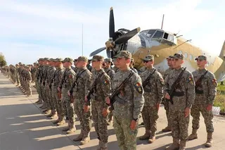 Soldați în uniforme de camuflaj stau în rând, cu fusiliile în față, în fața unui avion de propulsie vechi.