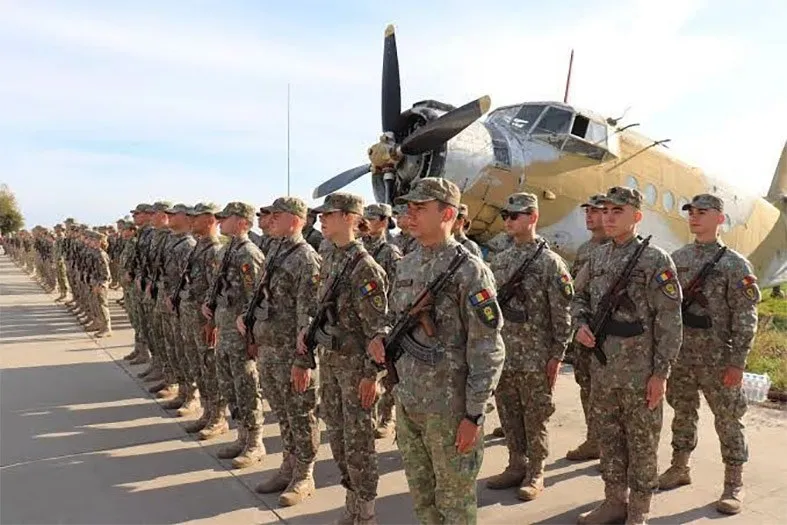 Soldați în uniforme de camuflaj stau în rând, cu fusiliile în față, în fața unui avion de propulsie vechi.