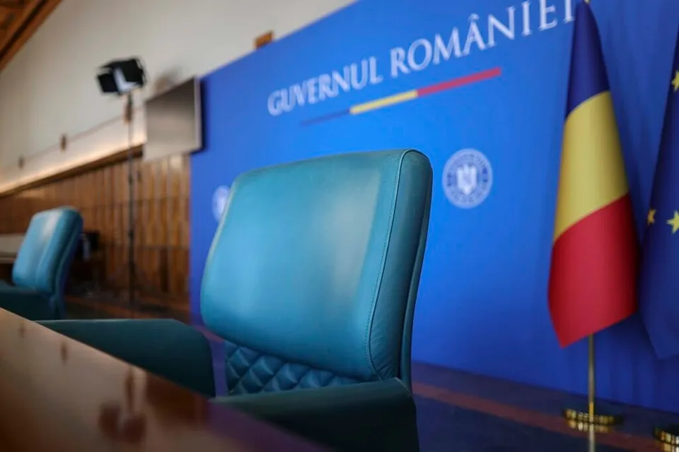 Două scaune de birou de culoare albastru sunt așezate în fața unui panou albastru pe care scrie Guvernul României lângă un steag.