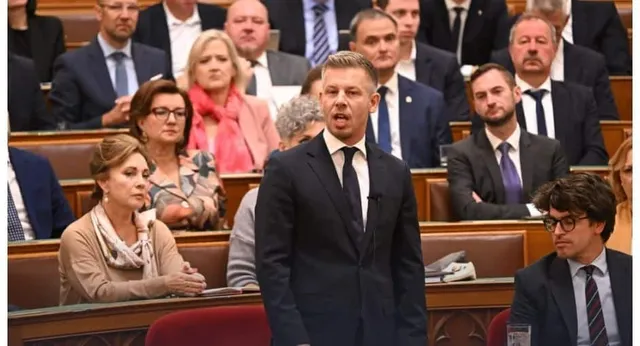 Un bărbat într-un costum închis și cravată neagră stă în picioare vorbind în fața unei grupări de oameni așezați pe scaune de lemn.