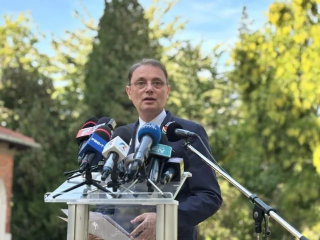 Un bărbat cu ochelari și costum închis stă în spatele unui podium cu mai multe microfoane de presă în fața unor copaci verzi.