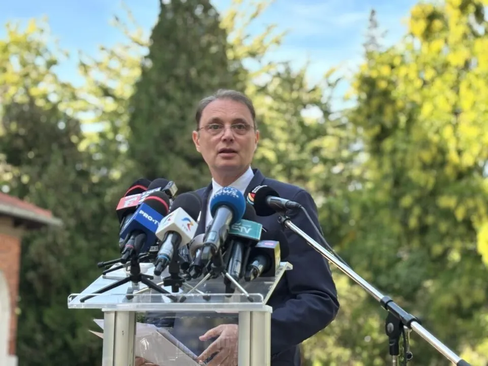 Un bărbat cu ochelari și costum închis stă în spatele unui podium cu mai multe microfoane de presă în fața unor copaci verzi.