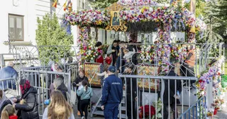 Oamenii stau în fața unei structuri metalice decorate cu flori colorate, lângă o icoană și niște tablouri religioase.