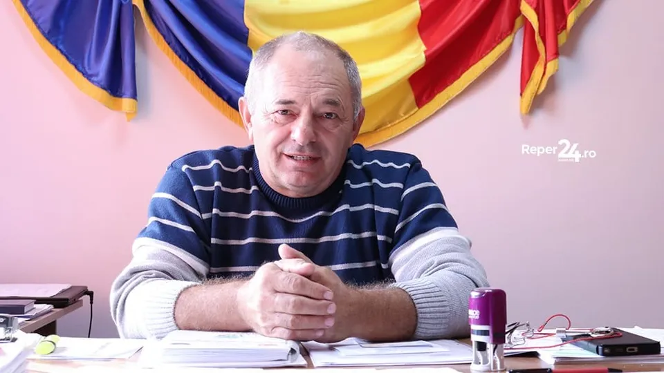Un bărbat cu tricou cu dungi orizontale albastre și albe stă la o masă cu hărți și obiecte de birou în față.