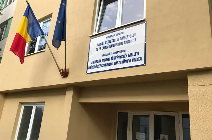 Un semn pe o clădire prezintă text în două limbi, lângă două steaguri suspendate pe o fațadă de culoare galben deschis.