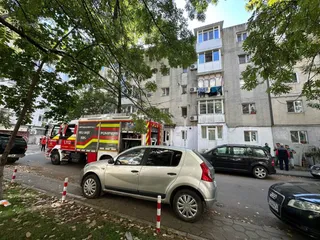 Un camion de pompieri este parcat pe o stradă lângă un bloc de locuințe, în fața căruia se află mai multe mașini și un grup de persoane.