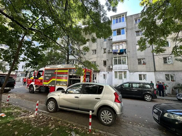 Un camion de pompieri este parcat pe o stradă lângă un bloc de locuințe, în fața căruia se află mai multe mașini și un grup de persoane.