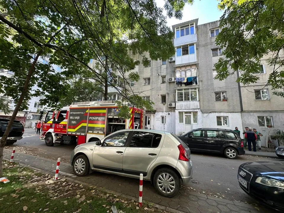 Un camion de pompieri este parcat pe o stradă lângă un bloc de locuințe, în fața căruia se află mai multe mașini și un grup de persoane.