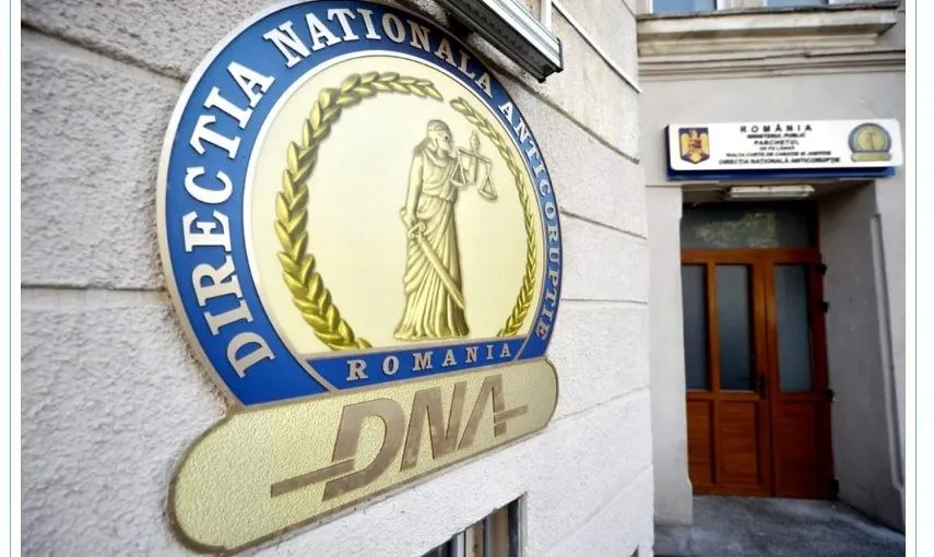 Un semn circular albastru și auriu cu inscripția Direcția Națională Anticorupție este montat pe o fațadă de clădire lângă o intrare cu uși de lemn.