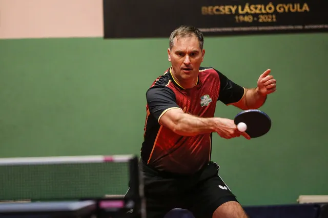Un bărbat cu tricou roșu și negru ține o paletă de tenis de masă și o minge albă deasupra unei mese de joc.
