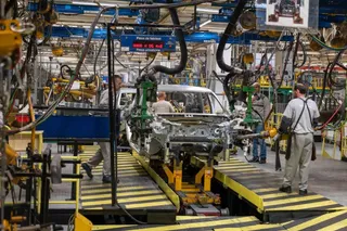 Muncitori asamblează o mașină pe o linie de producție într-o fabrică, cu diverse echipamente industriale și cabluri suspendate.