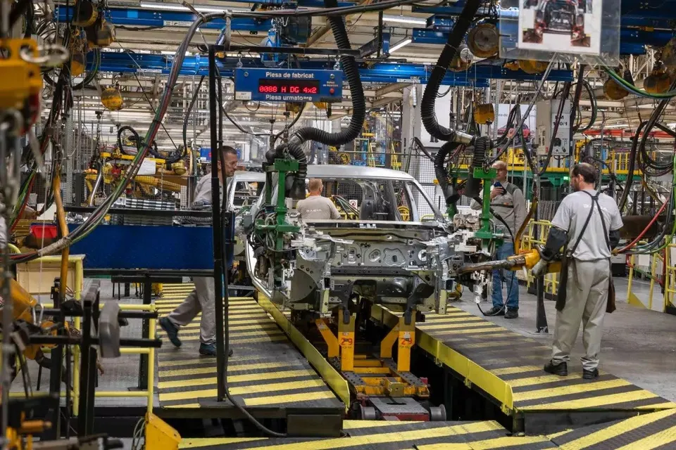 Muncitori asamblează o mașină pe o linie de producție într-o fabrică, cu diverse echipamente industriale și cabluri suspendate.