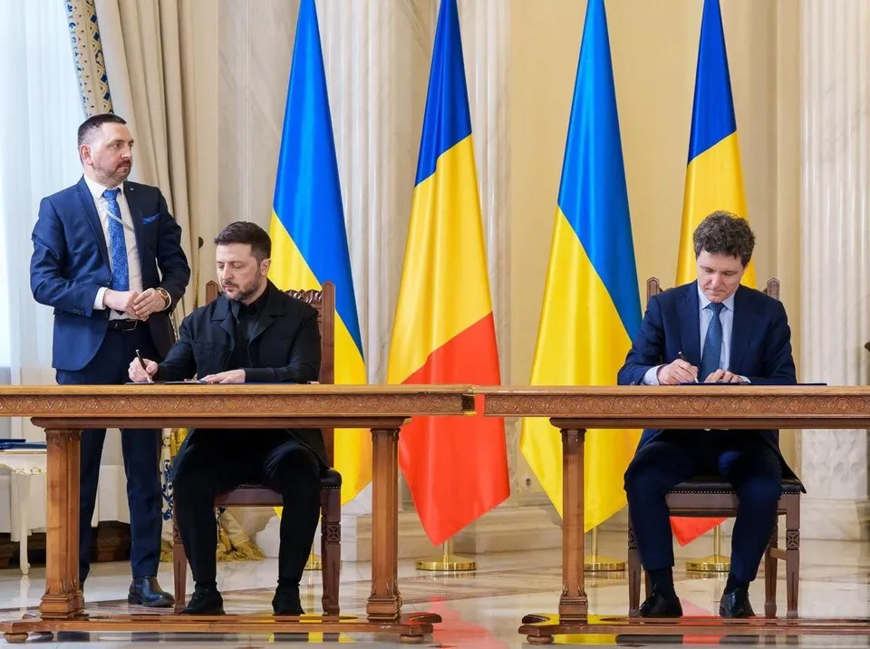 Trei bărbați în costume sunt prezenți într-o încăpere cu trei steaguri, doi dintre aceștia fiind așezați la o masă de lemn.