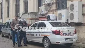 Un bărbat și o femeie stau lângă o mașină albă marcată cu inscripția POLITIA în fața unei clădiri cu aparate de aer condiționat.