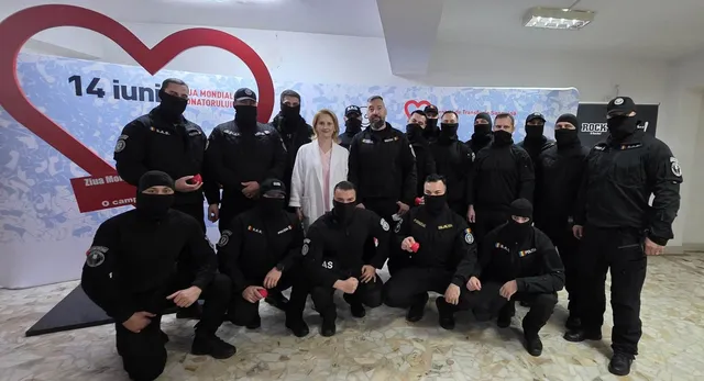 Un grup de persoane îmbrăcate în negru, cu măști de tip balaclavă, pozând alături de o femeie în alb, în fața unui banner cu un inimă roșie.