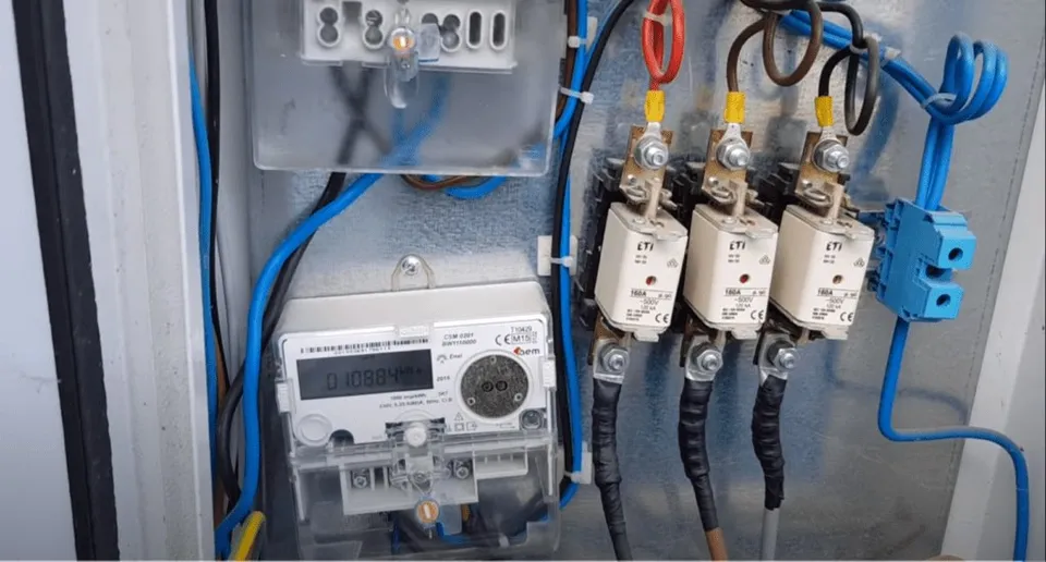 Un contor electric alb este conectat la cabluri colorate și la trei dispozitive de protecție montate pe o suprafață gri.