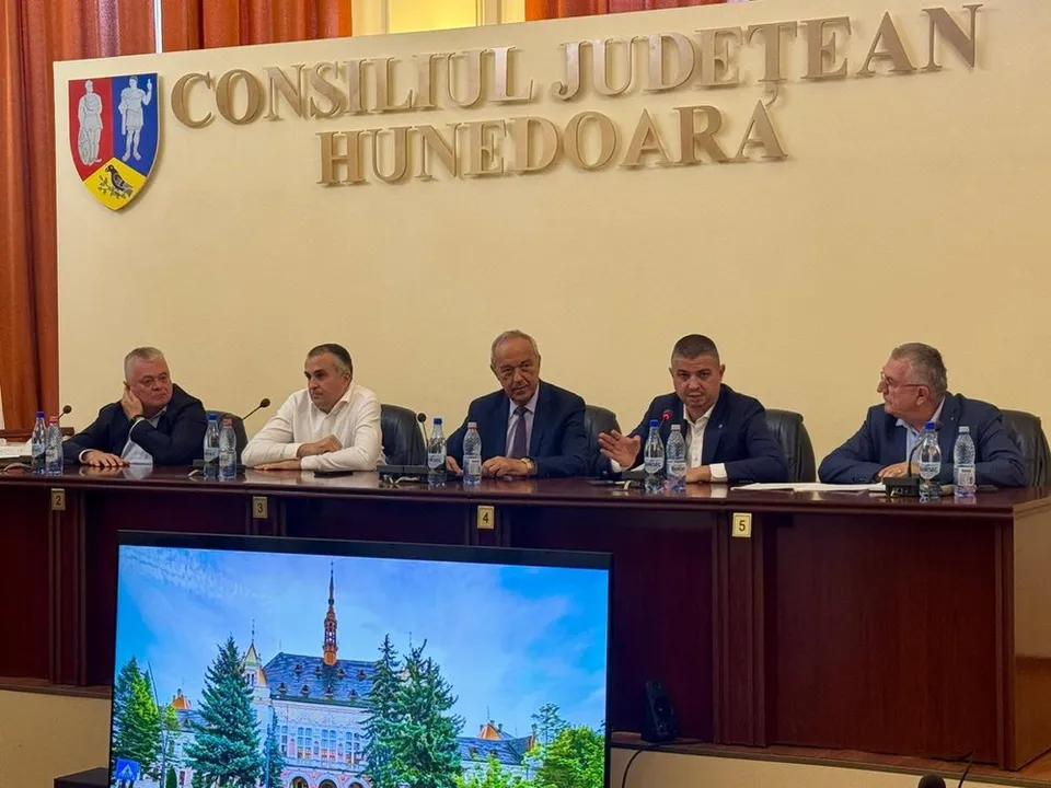 Șase bărbați stau așezați la o masă lungă de lemn în fața unui perete cu inscripții aurii și un blazon colorat.