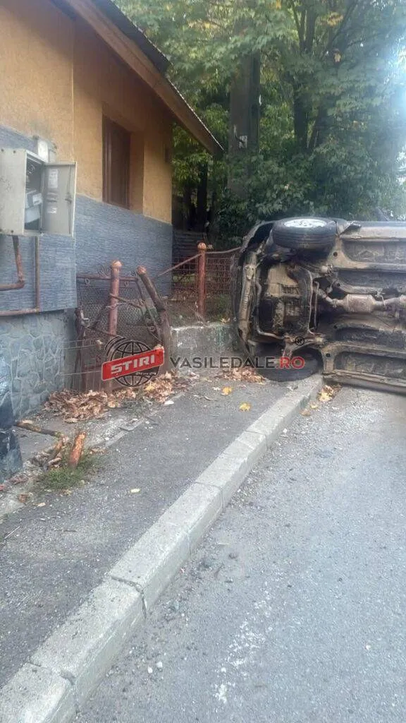 Un autoturism este răsturnat pe o parte lângă un gard metalic și o clădire cu un panou electric exterior.