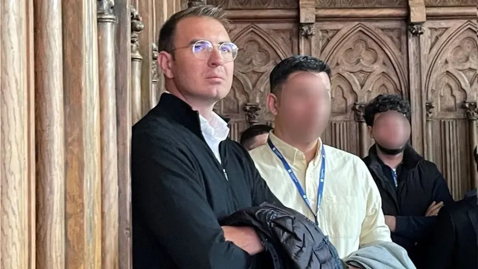 Un bărbat cu ochelari și sacou negru stă în fața unei panouri de lemn sculptat alături de alți doi bărbați.