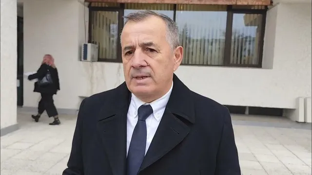 Un bărbat cu părul scurt, purtând o sacoșă neagră, o cămașă albă și o cravată albastră sub un palton negru, stă în fața unei clădiri.
