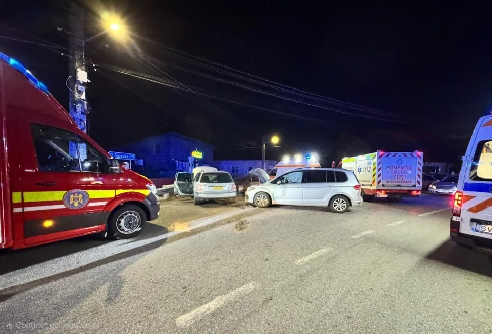 Două ambulanțe și mai multe autoturisme sunt parcate pe un drum asfaltat sub iluminat nocturn.