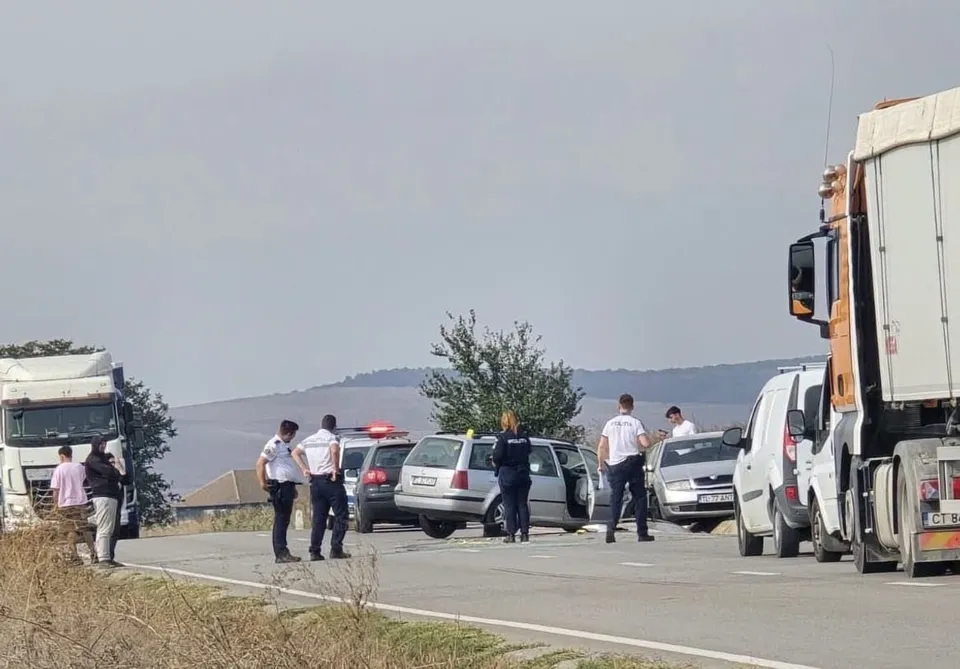 Mai mulți bărbați stau pe o porțiune de drum lângă două autoturisme și un camion sub un cer senin.