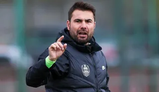 Fostul fotbalist Adrian Mutu, îmbrăcat într-o vestă neagră, vorbește și indică în timpul unei activități pe un teren sportiv.