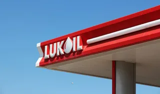 Un panou roșu cu inscripția LUKOIL albă este montat sub un acoperiș de benzinărie sub un cer albastru.