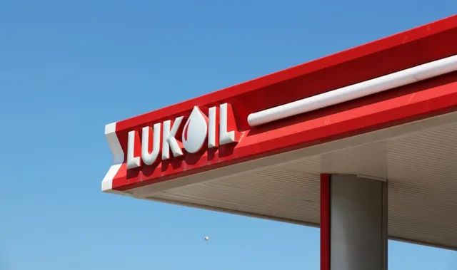 Un panou roșu cu inscripția LUKOIL albă este montat sub un acoperiș de benzinărie sub un cer albastru.