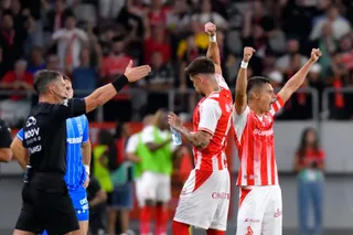 Trei jucători de fotbal în echipament alb și roșu sărbătoresc cu brațele ridicate pe un teren de sport, în fața unui arbitru și a unui public.