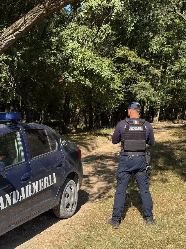 Un agent de jandarm stă lângă o mașină de serviciu albastră pe un drum de pământ într-o zonă cu copaci.