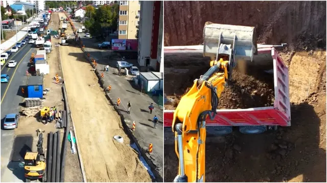 O excavatoare galbenă încarcă pământ într-un camion roșu, în timp ce în partea stângă se observă un șantier cu utilaje și mașini pe un drum.