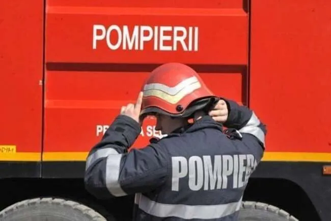 Un pompier cu cască roșie și uniformă închisă se află în fața unui vehicul de serviciu de culoare roșie pe care scrie POMPIERII.