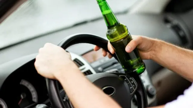 O persoană ține un volan cu o mână și o sticlă de sticlă verde cu lichid cu cealaltă mână în interiorul unui autovehicul.