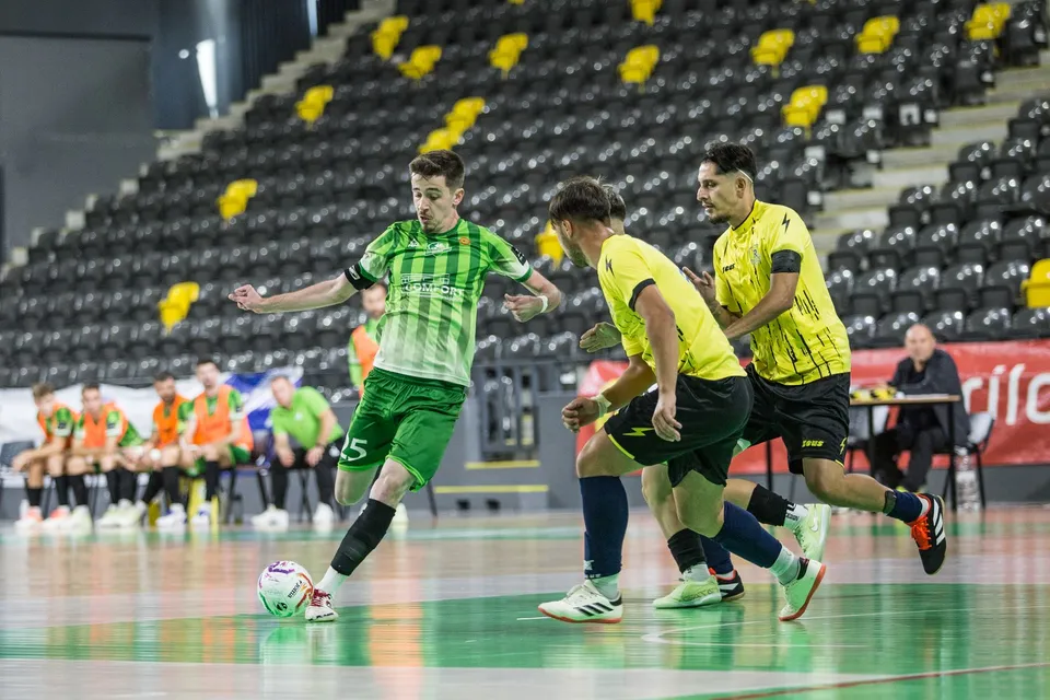 Un jucător în echipament verde și albastru lovește o minge pe un teren de fotbal de sală în fața unor adversari în echipament galben.