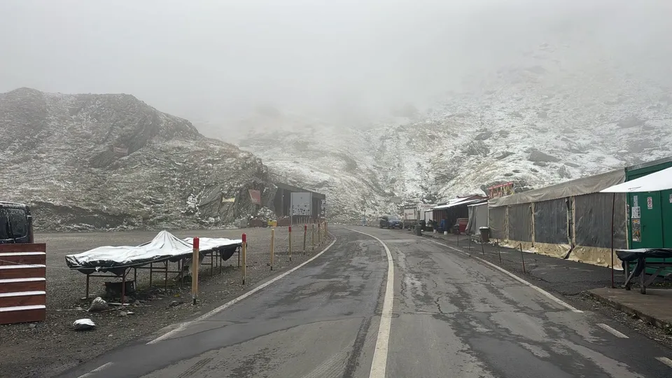Un drum asfaltat cu linii albe traversează un peisaj montan acoperit de zăpadă sub un cer înnorat și brumat.